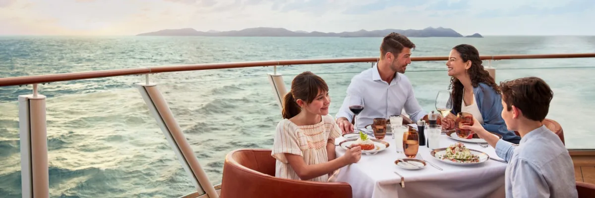 Familienaktivitäten bei NCL Eine vierköpfige Familie sitzt an einem Tisch an der Waterfront auf der Norwegian Encore und genießt ein Abendessen im Sonnenuntergang, während das Meer hinter Ihnen vorbeizieht.