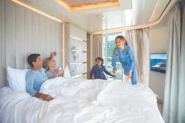 Familienkabinen bei NCL Eine vierköpfige Familie in einer Familienkabine im The Haven Bereich der Norwegian Encore. Die beiden Eltern liegen schon unter der Decke und die Tochter springt gerade auf das Bett.