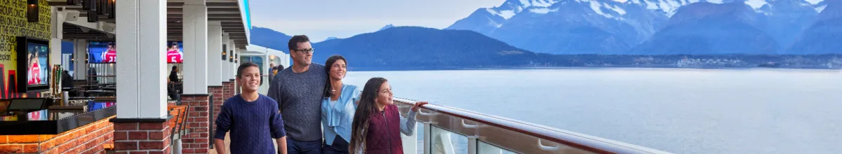 Eine Familie blickt von der Reling des weitläufigen Außendecks der Norwegian Cruise Line Lifestyle-Flotte auf eine majestätische Gebirgslandschaft mit Gletschern.