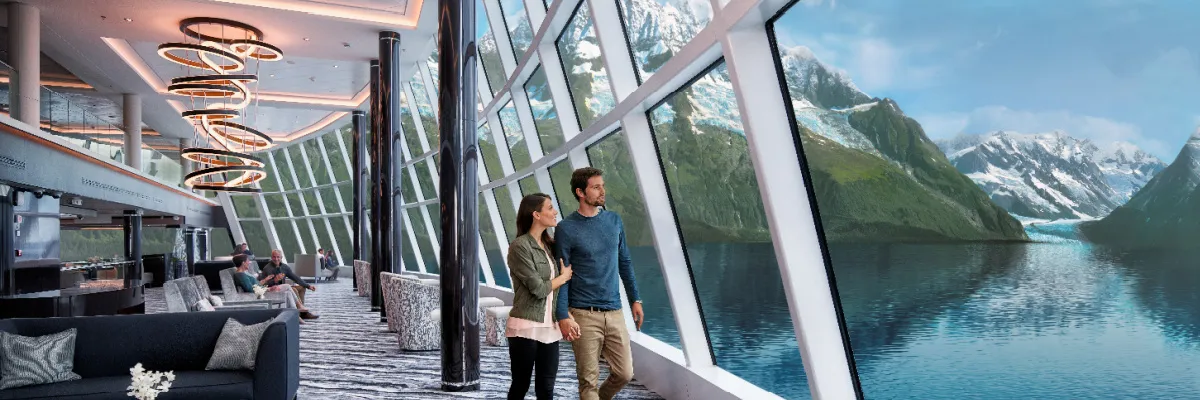 NCL Lifestyle-Flotte: Grenzenlose Aussichten in der Observation Lounge Ein Paar blickt aus den bodentiefen Panoramafenstern der luxuriösen Observation Lounge der Norwegian Cruise Line Lifestyle-Flotte auf eine spektakuläre Fjordlandschaft mit Gletschern.