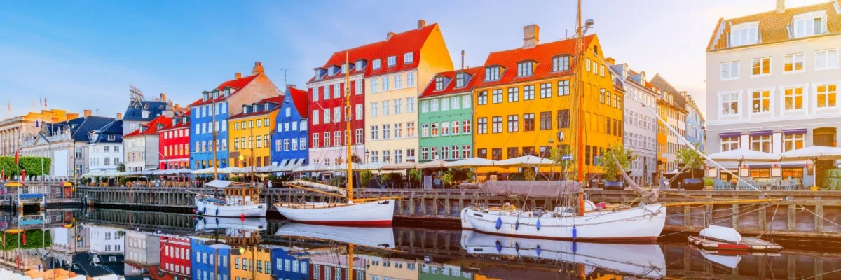 Kabinenupgrades für Ihre Kreuzfahrt durch Nordeuropa Bunte Häuser am Nyhavn in Kopenhagen, die sich an einem sonnigen Tag im Wasser spiegeln. Auf dem Wasser vor der Häuserfront sind verschiedene Segelboote zu sehen.