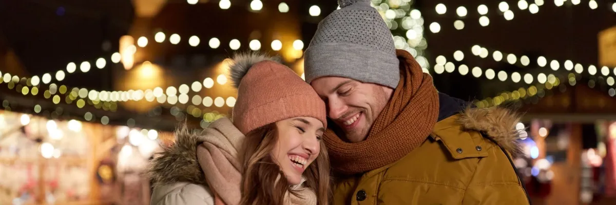 Kreuzfahrt für gute Laune entdecken Ein Pärchen trägt Wintermützen, lächelt und im Hintergrund sind Lichter zu sehen.