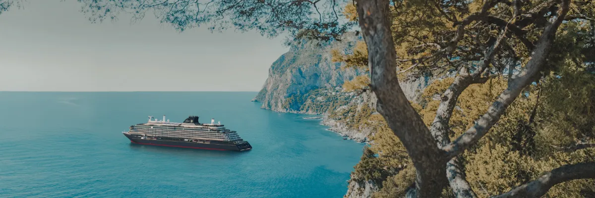 Eines der EXPLORA Schiffe im Westlichen Mittelmeer vor einer Klippe am Meer, die mit Bäumen bewachsen ist.