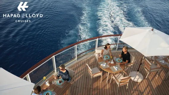 Exklusiver Genuss an Deck – Hapag-Lloyd Cruises Club Vorteilswelt Gäste beim exklusiven Freiluft-Dining auf dem Holzdeck eines Hapag-Lloyd Cruises Luxusschiffs mit Blick auf das Meer.
