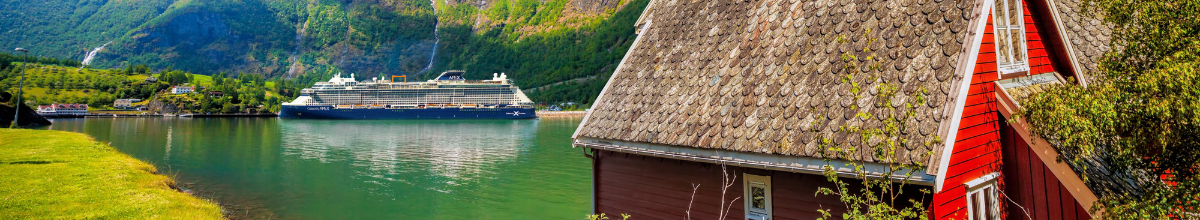Die Celebrity Apex, ein Schiff von Celebrity Cruises liegt an einem Hafen in Norwegen. Im Vordergrund steht ein typisch norwegisches, rotes Haus.