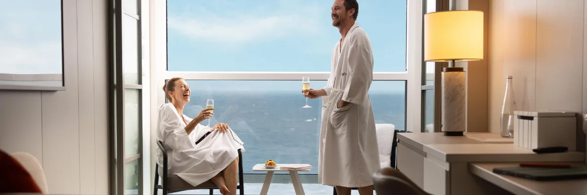Kabinen auf Celebrity Apex Ein Paar steht in Bademänteln in Ihrer Infinity Veranda Kabine und genießt einen Champagner neben den Panoramafenstern.