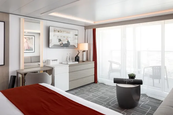 Kabinen auf Celebrity Beyond Eine helle Kabine auf der Celebrity Beyond mit roten Akzenten und einer großen Panorama-Glasfront, die zum Balkon führt.