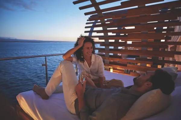 Abend im Außenbereich der Celebrity Flora Paar mit Sektgläsern auf einem Außenbett an Deck der Celebrity Flora in der Abenddämmerung.