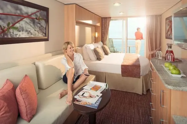 Kabinen der Celebrity Constellation Einne Frau sitzt auf dem Sofa der Prime Aquaclass Balkonkabine auf Celebrity Constellation. Ihr Mann steht auf dem Balkon und schaut auf das Meer.