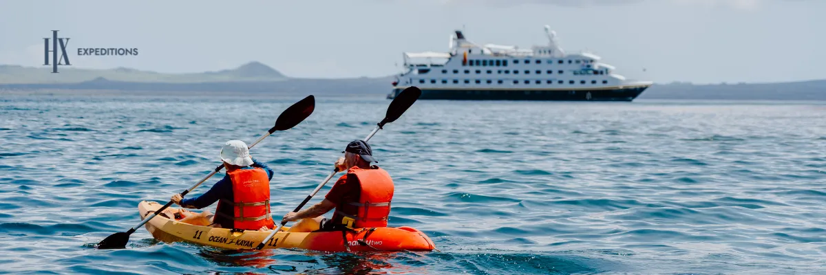 Expeditionskreuzfahrten mit HX Zwei Personen fahren an der Küste der Rabida Insel bei den Galápagos Inseln ein Kayak. In Hintergrund ankert das Schiff MS Santa Cruz II.