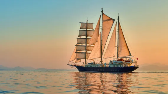 Segelkreuzfahrt buchen Ein großes Segelschiff mit drei Masten segelt entspannt mit geöffneten Segeln über das ruhige Wasser.