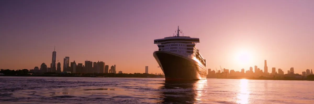 Mit den Cunard Queens im Sommer 2027 über den Atlantik. Frontansicht der Queen Mary 2 vor der Skyline von New York in der Abenddämmerung.