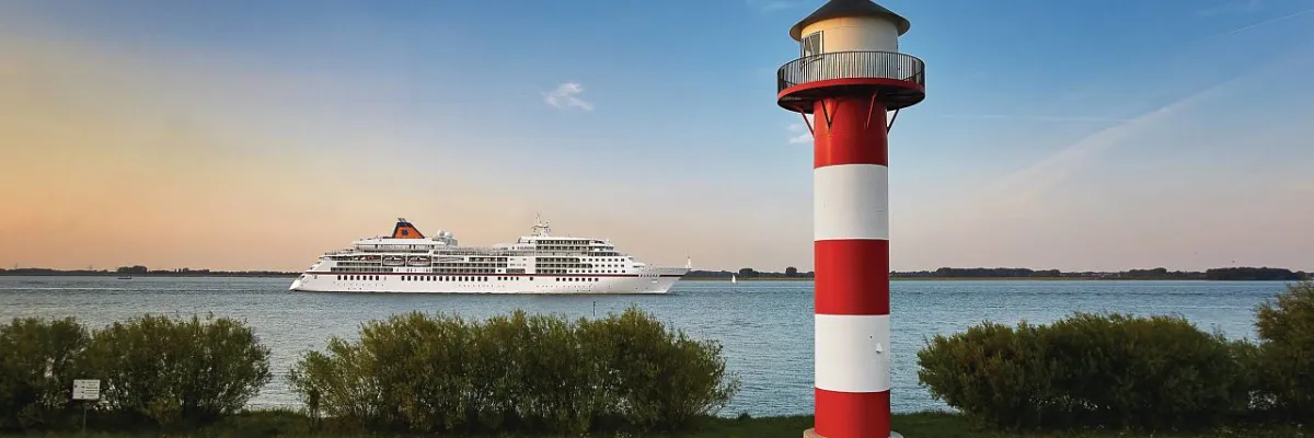 Das Luxusschiff EUROPA zieht auf ihrer Reise Parks & Gardens stilvoll an einem rot-weiß gestreiften Leuchtturm vorbei Die EUROPA von Hapag-Lloyd Cruises gleitet bei sonnigem Wetter an einer grünen Küstenlandschaft mit Leuchtturm entlang.