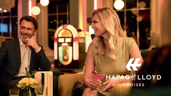 Eine elegante Dame mit Cocktail und ein Herr im Anzug genießen die Abendstimmung in der Lounge an Bord von Hapag-Lloyd Cruises.