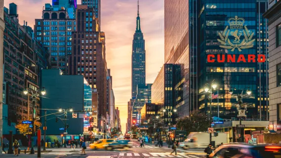 Blick entlang einer belebten Straße in Manhattan mit dem Empire State Building in der Abenddämmerung und dem goldenen Cunard Logo für das Arrangement The New Yorker.