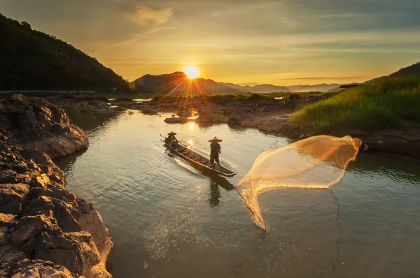 Mekongkreuzfahrt mit Emerald Cruises Ein Fischer auf einem Boot im Sonnenuntergang wirbelt Wasser mit seinem Paddel auf.