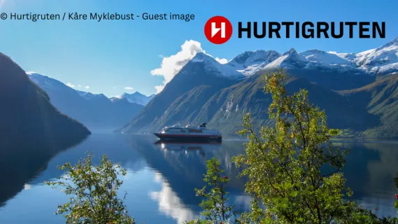 Bis zu 700 € Bordguthaben für Ihre Hurtigruten Kreuzfahrt 2027 oder 2028 Schiff der Hurtigruten in einer frühlingshaften Fjordlandschaft in Norwegen.
