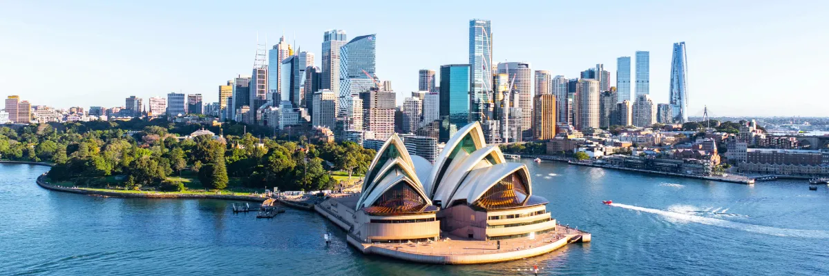 Australien & Neuseeland entdecken Ein wunderschöner Blick auf das Sydney Opera House mit glitzerndem Wasser, modernen Gebäuden und einem grünen Park im Hintergrund.