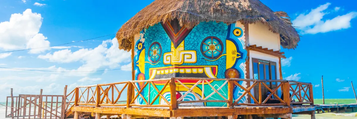 Karibische Impressionen und Kultur erleben mit Mein Schiff® Bunt bemaltes Strandhaus mit Reetdach auf einem Holzsteg über dem türkisblauen Meer – Karibik-Ausflug während einer <em>Mein Schiff</em>® Kreuzfahrt.