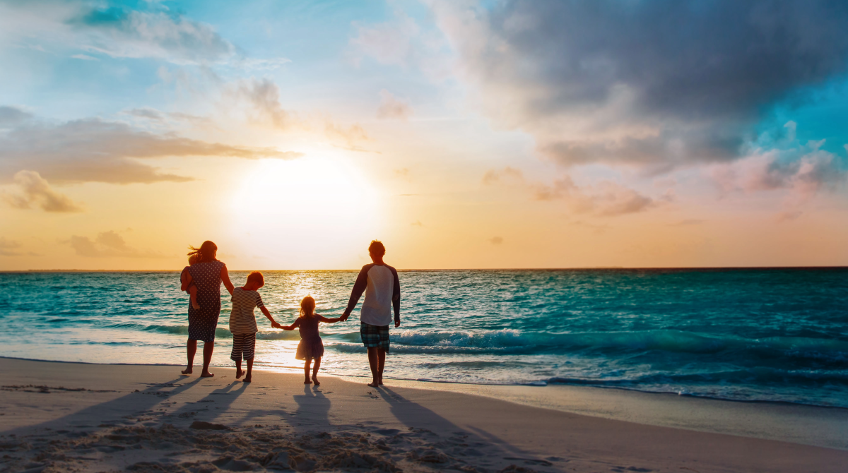 Eine Familie steht Hand in Hand am Strand und blickt auf das Meer – Symbol auf ein Familienurlaub.