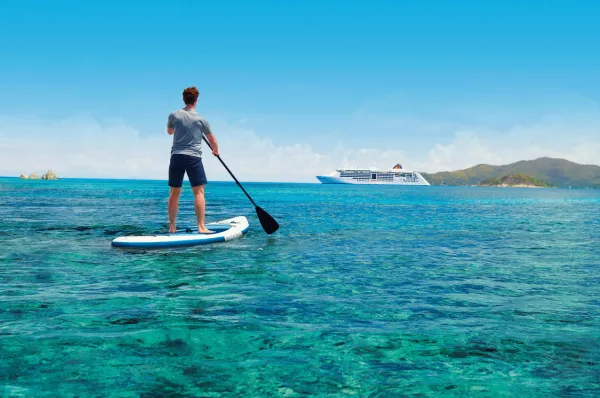 Ein Mann steht auf einem SUP im karibischen Meer und schaut auf das Hapag-Lloyd Schiff, dass besondere Einzelreisenden-Konditionen für ihn angeboten hat.