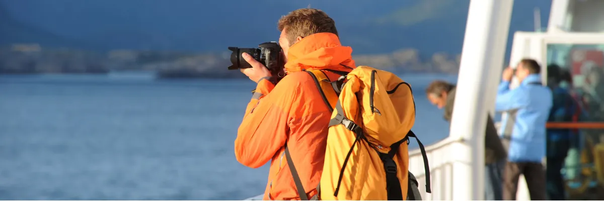 Alleine auf Kreuzfahrt reisen Ein Mann in wetterfester Kleidung und mit Rucksack steht an Deck bei Hurtigruten und fotografiert die Landschaft.