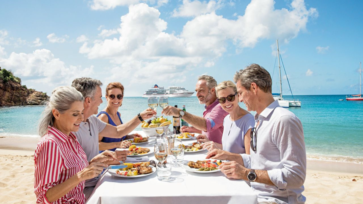 Eine Gruppe von Gästen genießt ein exklusives Gourmet-Lunch direkt am Sandstrand vor türkisfarbenem Meer, während im Hintergrund das Luxusschiff EUROPA 2 vor Anker liegt.