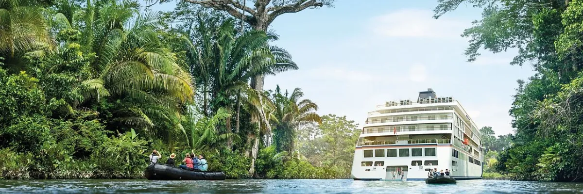Hapag-Lloyd Cruises Tropen-Expedition: Gäste erkunden Flusslandschaften im Zodiac Gäste in einem Zodiac-Schlauchboot erkunden dichten, exotischen Regenwald entlang eines tropischen Flusses, während im Hintergrund ein Expeditions-Schiff der HANSEATIC-Klasse ankert.