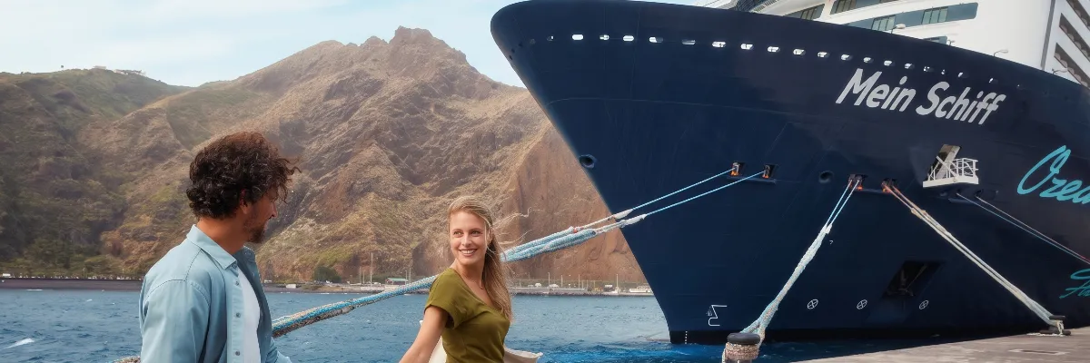 Gäste vor dem Bug eines TUI Schiffes an der Kaimauer vor bergiger Küstenlandschaft Zwei Kreuzfahrtgäste spazieren lächelnd an der Pier entlang, während im Hintergrund der markante blaue Bug eines TUI Schiffes vor einer imposanten Bergkulisse festgemacht ist.
