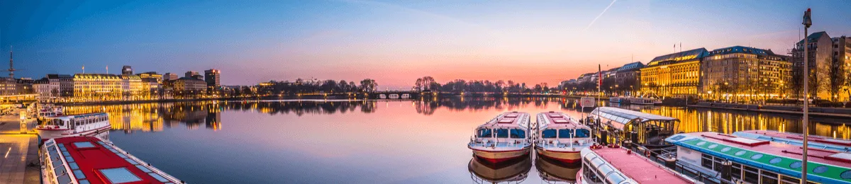 Panoramaaufnahme der Binnenalster in Hamburg bei Sonnenuntergang. Im Vordergrund liegen Alsterdampfer am Anleger des Jungfernstiegs, während sich die beleuchteten Gebäude der Innenstadt im ruhigen Wasser spiegeln.