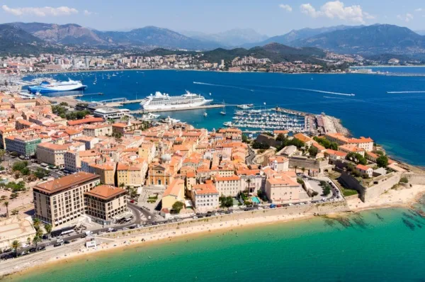 Ein Kreuzfahrtschiff ankert vor Ajaccio am Hafen. Die Stadt am Meer mit kleinen ockerfarbenen Häusern ist zu sehen.