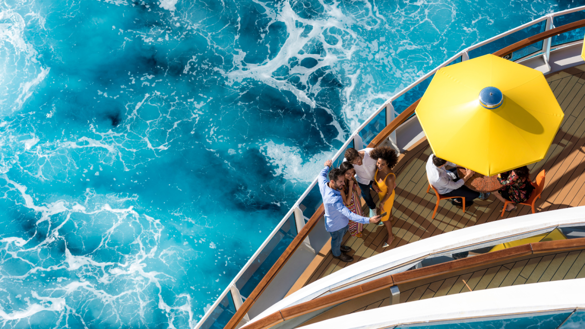 Gäste verweilen auf dem Holzdeck eines Costa-Kreuzfahrtschiffs unter gelben Sonnenschirmen mit Blick auf das türkisfarbene Meer.