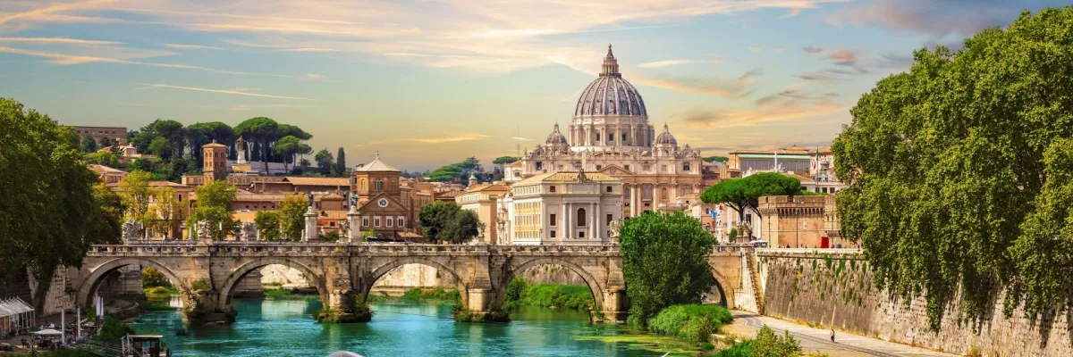 Brücke über den Tiber in Rom mit dahinter liegendem Petersdom. Am Ufer des Flusses sind üppig grüne Bäume zu sehen.