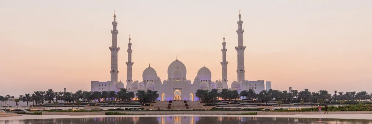 Frontansicht der imposanten Scheich-Zayid-Moschee in Abu Dhabi im Licht des beginnenden Sonnenuntergangs. Vor der Moschee sind viele Palmen zu sehen, davor erstreckt sich ein See, in dem sich das Gebäude spiegelt.