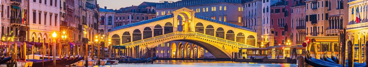 Die beleuchtete Rialtobrücke über den Canal Grande in Venedig zur blauen Stunde mit am Ufer vertäuten Gondeln im warmen Laternenlicht.