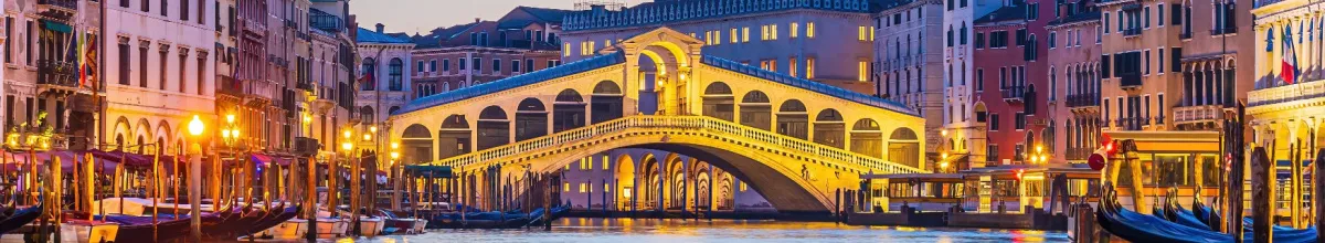 Die beleuchtete Rialtobrücke über den Canal Grande in Venedig zur blauen Stunde mit am Ufer vertäuten Gondeln im warmen Laternenlicht.