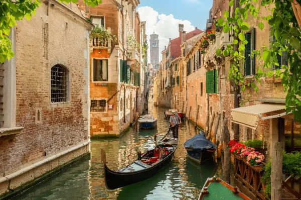 Eine traditionelle schwarze Gondel mit Gondoliere in einem malerischen, von historischen Backsteinhäusern gesäumten Seitenkanal in Venedig mit Blick auf einen fernen Glockenturm.