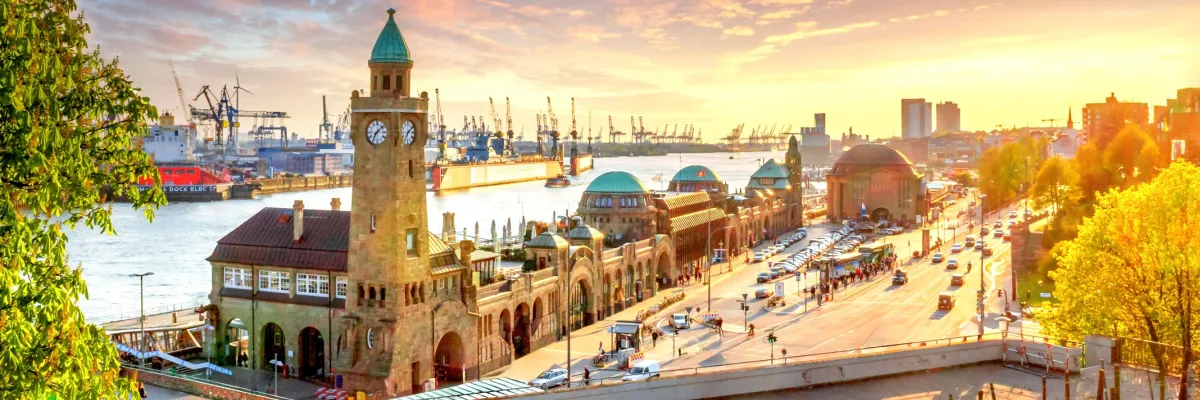 Panorama-Blick auf die Hamburger Landungsbrücken bei goldenem Sonnenuntergang mit dem Hafenbecken und Kränen im Hintergrund.