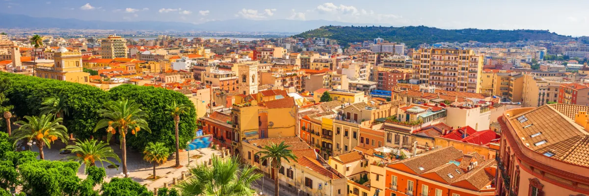 Panoramablick über die sonnendurchfluteten Dächer von Cagliari auf Sardinien mit Palmen im Vordergrund und mediterraner Architektur unter blauem Himmel.