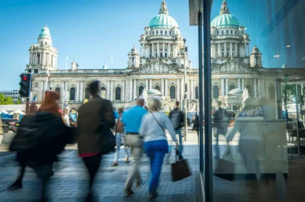 Viele Menschen mit Einkaufstaschen gehen auf der Oberstraße von Belfast, wobei im Hintergrund die Beltfast City Hall zu sehen ist