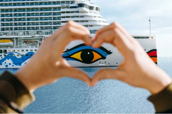Zwei Hände formen ein Herz vor dem ikonischen Auge am Bug von AIDAcosma. Die perspektivische Aufnahme vor azurblauem Wasser symbolisiert Urlaubsglück und die emotionale Vorfreude auf eine spontane Auszeit auf See.