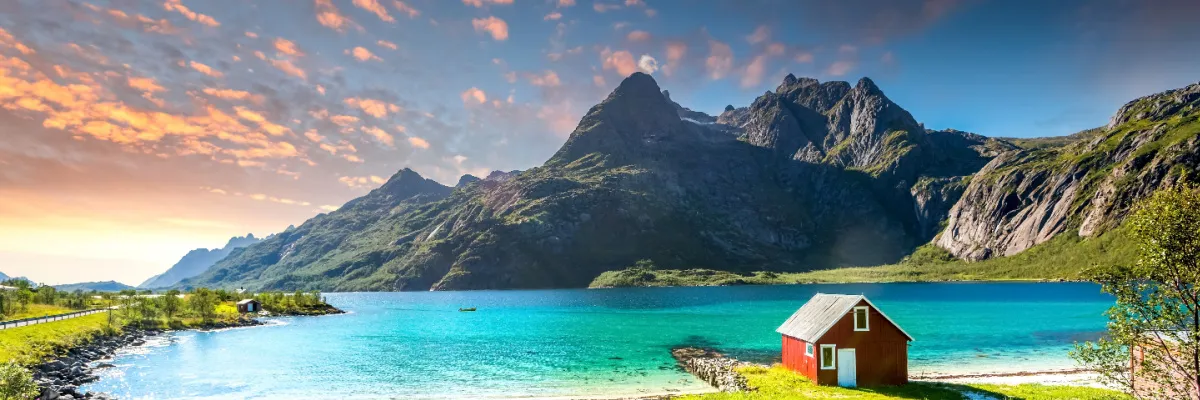 Malerische rote Holzhütte an einer türkisfarbenen Bucht in den norwegischen Fjorden vor einer imposanten Bergkulisse. Ein Sinnbild für die skandinavische Naturidylle, die Reisende auf den Nordeuropa-Routen ab Hamburg oder Kiel erwartet.
