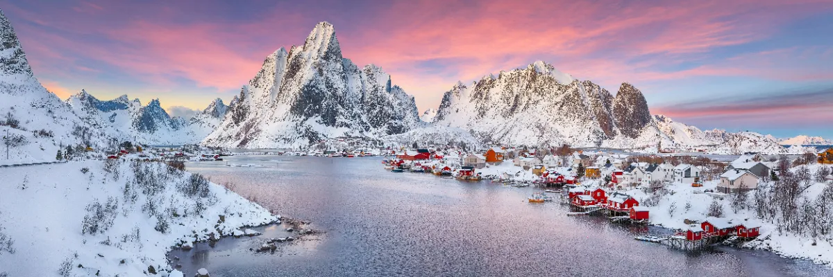Panoramaaufnahme eines norwegischen Küstenortes mit roten Fischerhäusern vor massiven, schneebedeckten Berggipfeln während der Dämmerung mit rosa-leuchtendem Himmel.
