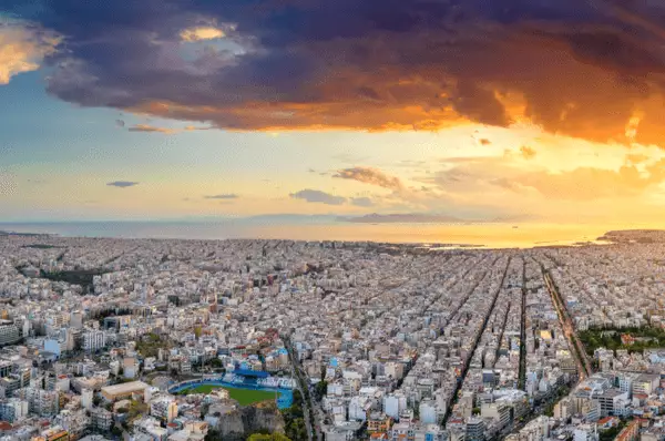 Panoramablick über Athen. Am Himmel lugt die untergehende Sonne unter einer dunklen Wolke hervor und wirft ihre Strahlen auf die Stadt.