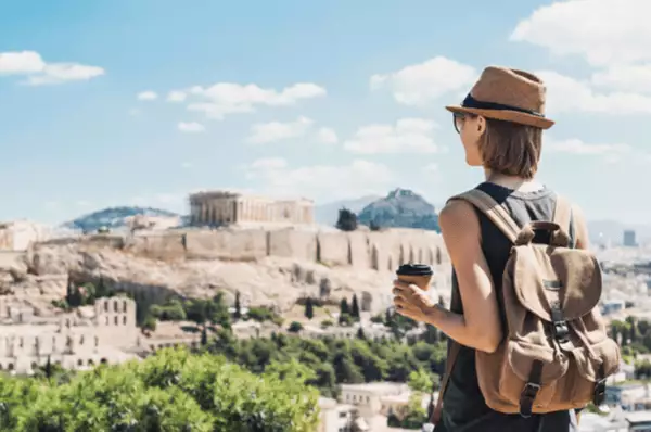 Eine junge Frau mit Rucksack und Sonnenbrille blickt von fern auf die Akropolis in Athen.