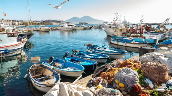 Kleine Fischerboote im Hafen von Neapel mit Netzen am Kai und dem Vulkan Vesuv im sonnigen Hintergrund – ideal für eine Kreuzfahrt nach Neapel.