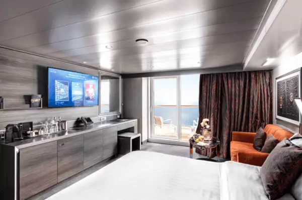 MSC Yacht Club Deluxe Suite auf der MSC Euribia Blick in die MSC Yacht Club Deluxe Suite mit Doppelbett, Schreibtisch, TV, Sessel und einem bestuhlten Balkon hinter einer Glastür.