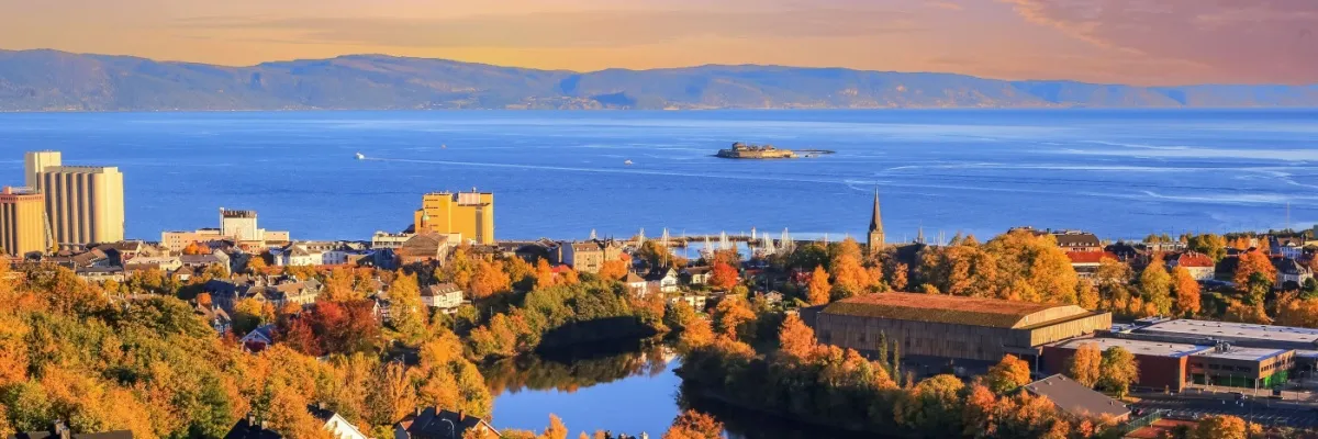 Blick über die Dächer von Trondheim auf den Fjord mit der Insel Munkholmen. Vorne die Häuser der Stadt mit vielen herbstlich-bunt gefärbten Bäumen, im Hintergrund das gegenüberliegende Fjordufer.