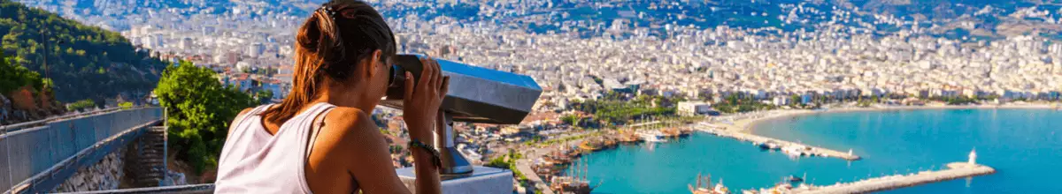 Eine junge brünette Frau blickt durch ein Fernglas an einem Aussichtspunkt auf den Hafen von Alanya, Türkei.