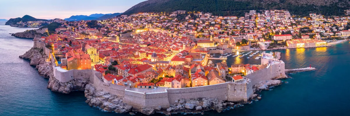 Dubrovnik am Abend – Stimmungsvoller Landausflug zur blauen Stunde Panorama-Blick auf die beleuchtete Altstadt von Dubrovnik während der Abenddämmerung mit der hell erleuchteten Stadtmauer und kleinen Booten im Hafen vor der kroatischen Küste.